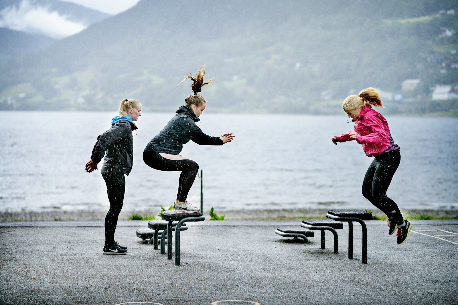 woman in pink jacket and black pants jumping on black bench near body of water during